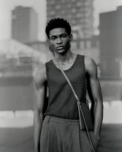 Monochrome portrait of a Black man wearing a textured knit tank top, pleated trousers, and a slim crossbody bag outdoors.