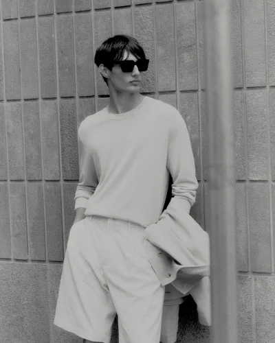 Black and white photo of a man wearing dark sunglasses, a light crewneck sweater, and tailored shorts against a textured tiled wall.