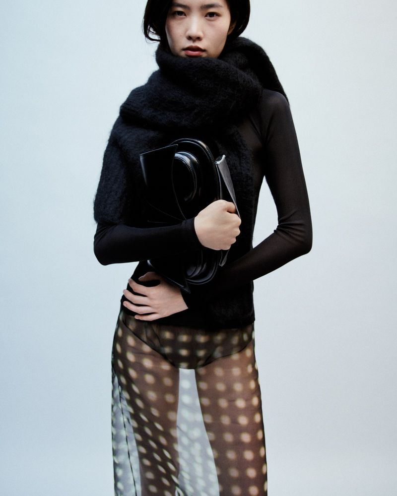 Model in black top and sheer dotted skirt holding a structured handbag against a plain background