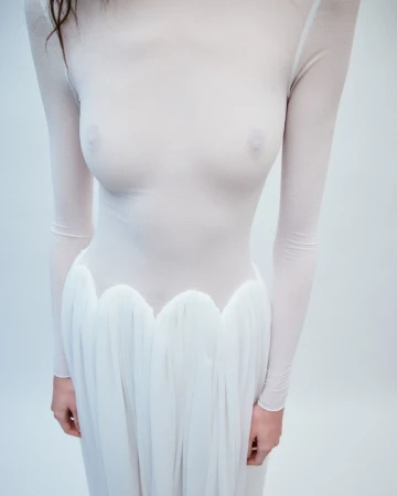 Model wearing a sheer long-sleeve white top and pleated skirt against a plain studio background.