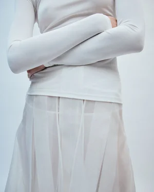 Model wearing a long-sleeved white top and sheer pleated skirt with arms crossed against a plain background.