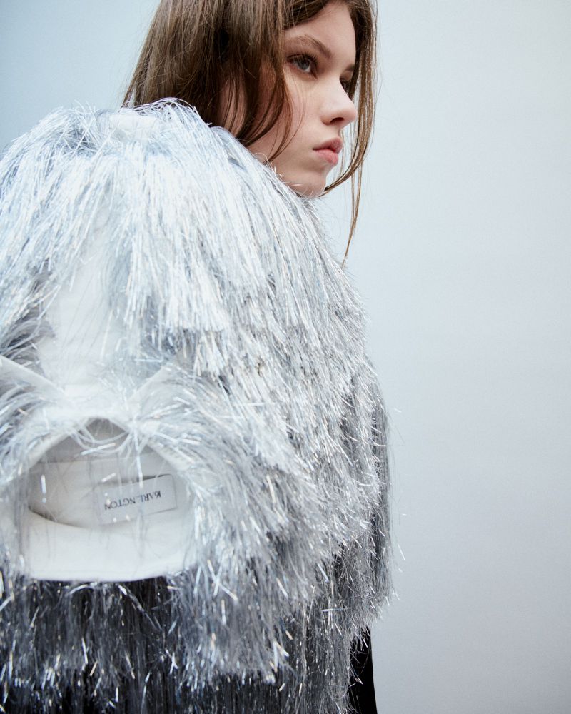 Woman in a silver fringe jacket stands in a studio with a pale background. This is a studio shot.