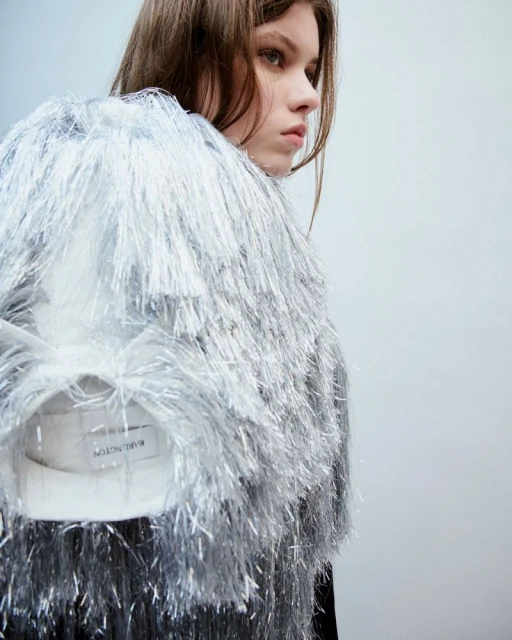 Woman in a silver fringe jacket stands in a studio with a pale background. This is a studio shot.