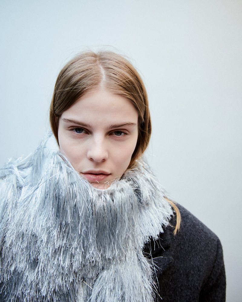 Close-up portrait of an adult female model with fair skin and light brown hair wearing a grey coat with a fringed silver shawl.