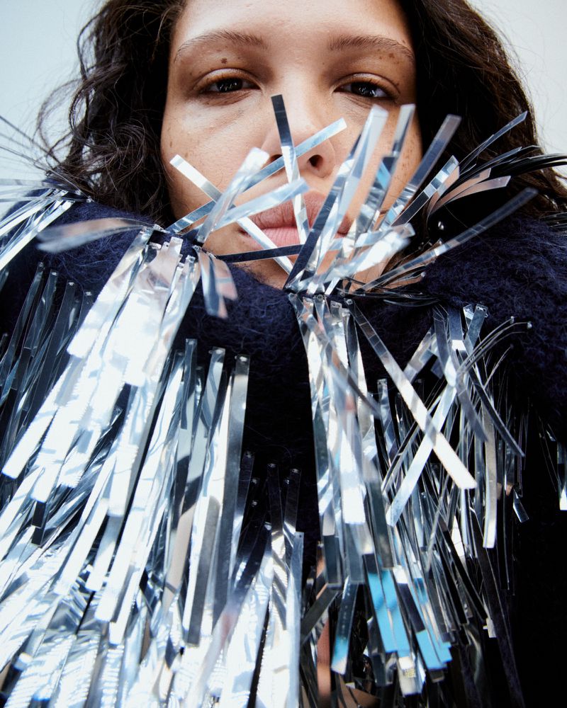 Close-up shot of a woman wearing a dark sweater with shiny silver fringe, highlighting fashion and studio lighting, indoor setting.