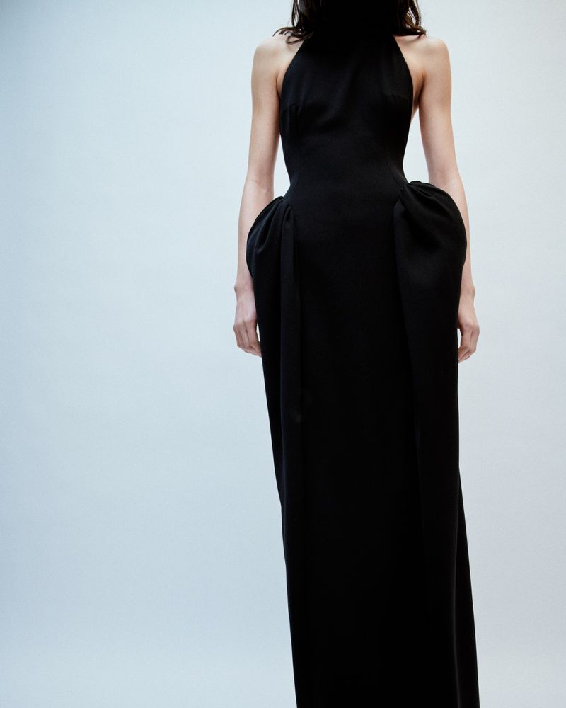Woman in elegant black halter gown with sculptural hip detail. Formal fashion shot against a pale background.