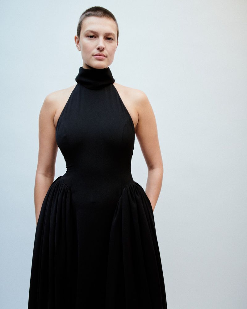 Studio shot of a woman with short hair wearing a sleeveless black dress with a high neckline and a full skirt, with neutral background.