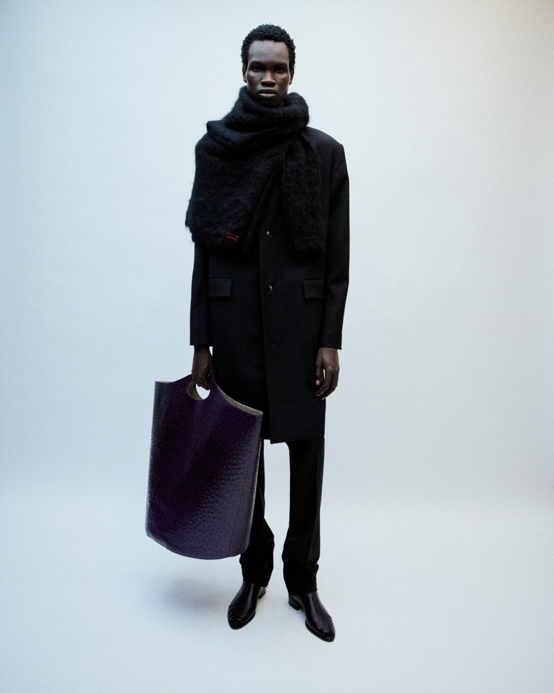 Model in black coat wearing a large textured scarf holding a deep purple tote bag against a neutral studio backdrop