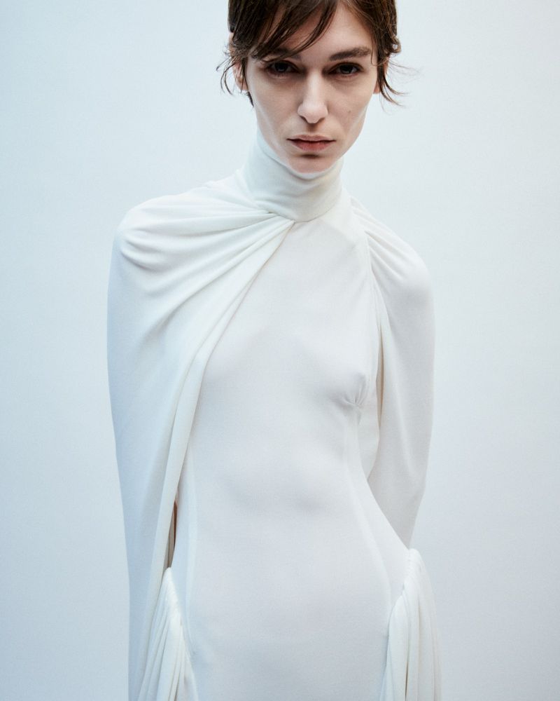 Indoor shot of a slender woman in an elegant white dress with a high neckline and draped details against a light background.