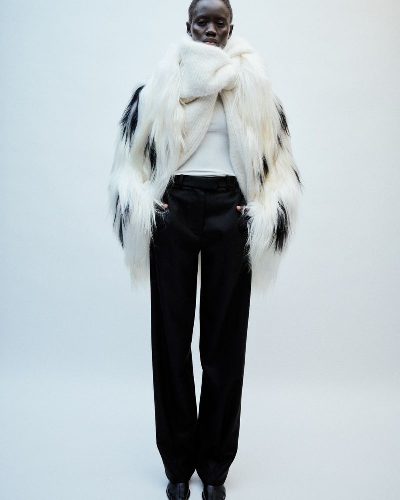 Dark-skinned model stands in a photo studio wearing a large black and white faux fur coat, white shirt, black pants, and black shoes.