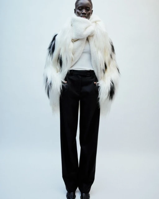 Dark-skinned model stands in a photo studio wearing a large black and white faux fur coat, white shirt, black pants, and black shoes.