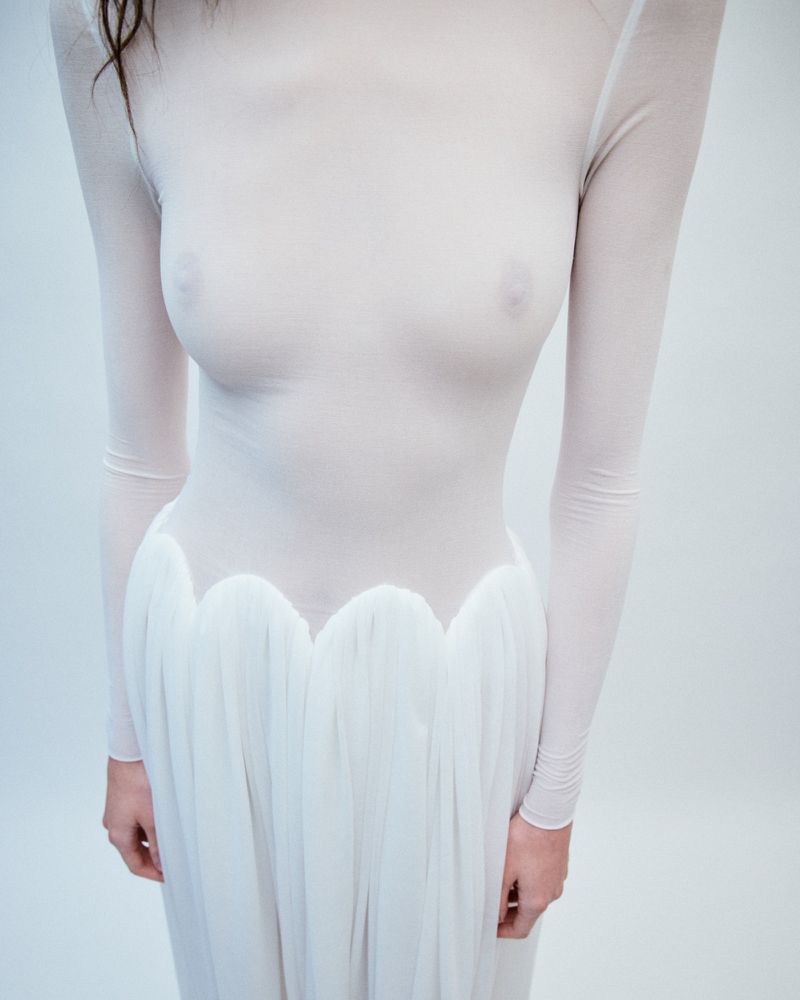 A woman in a sheer, long-sleeved white dress poses in a minimalist, white studio setting, showcasing the dress's delicate fabric and design.