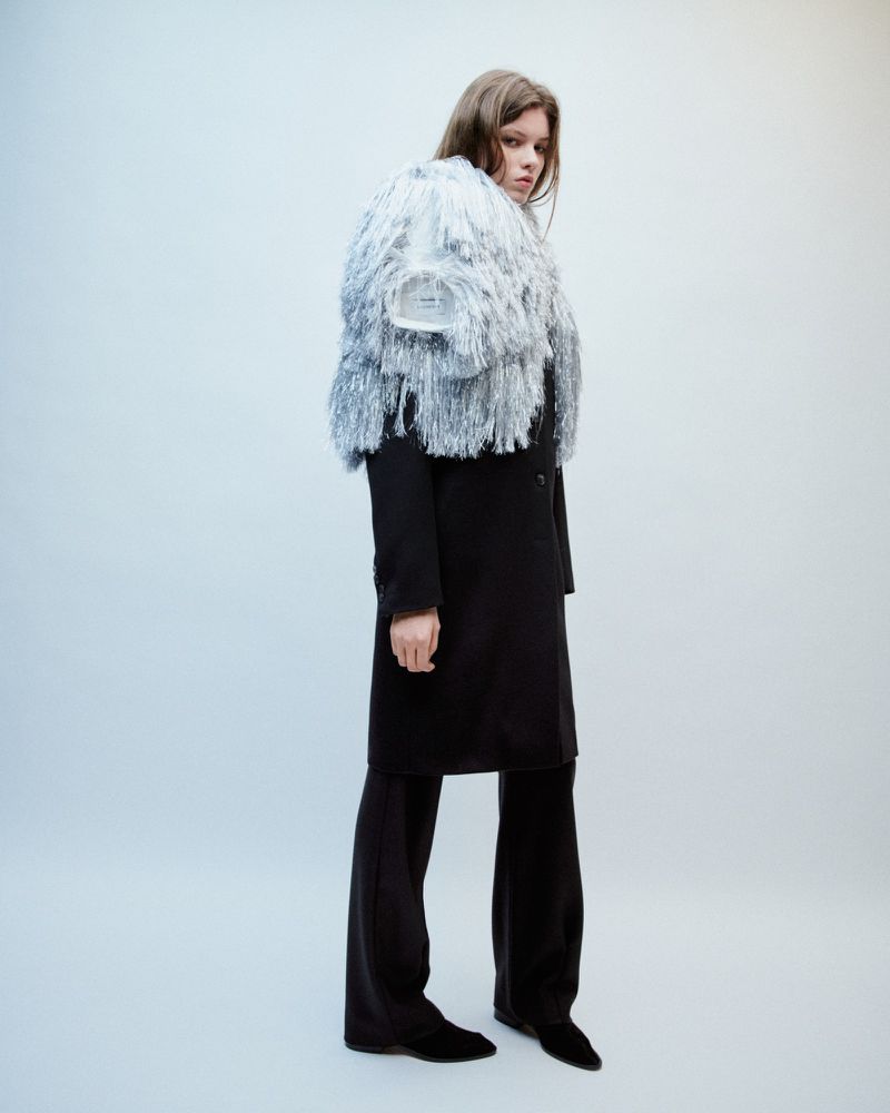 Full shot of a young woman wearing a black overcoat with fringe detail in a light studio. Fashion editorial portrait.