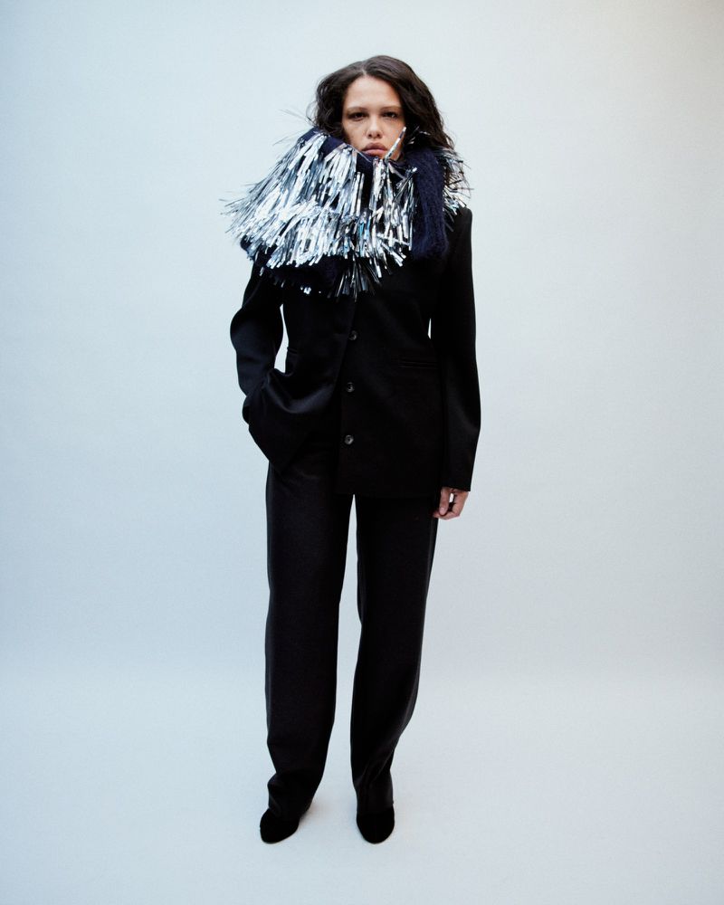 Woman with dark hair poses in a black suit with silver fringe detail against a white background in a fashion photography studio.