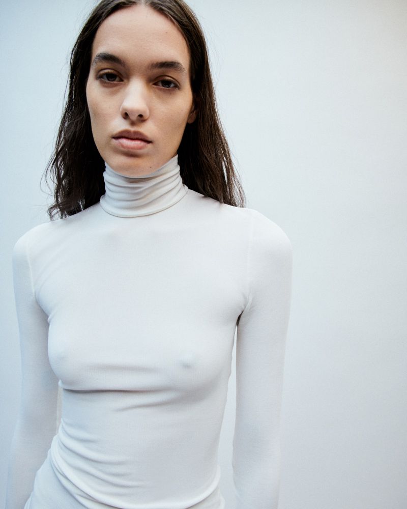 Close-up shot of a female adult wearing a white turtleneck shirt against a white background in a studio, showcasing fashion and beauty.
