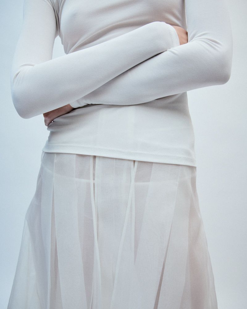 Model wearing a long-sleeved white top and sheer pleated skirt with arms crossed against a plain background.