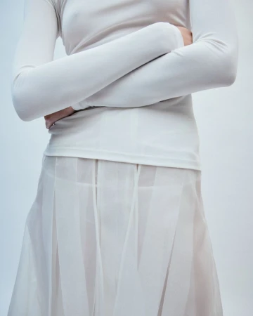 Model wearing a long-sleeved white top and sheer pleated skirt with arms crossed against a plain background.