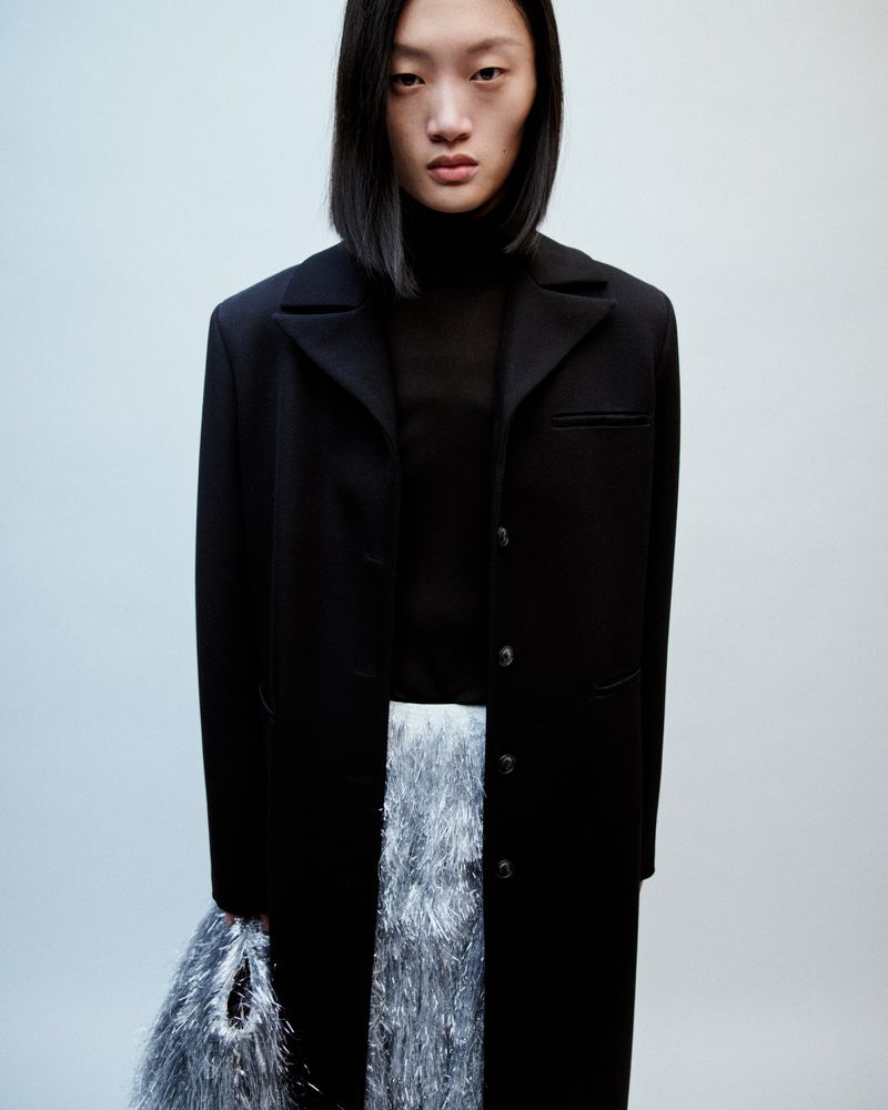 Fashion shot of a young Asian woman in a black coat and iridescent fringed skirt, standing in a studio with a neutral background.