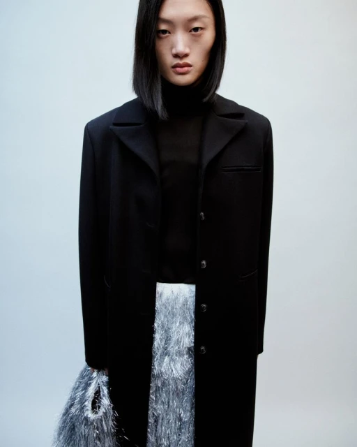 Fashion shot of a young Asian woman in a black coat and iridescent fringed skirt, standing in a studio with a neutral background.