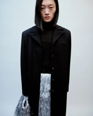 Fashion shot of a young Asian woman in a black coat and iridescent fringed skirt, standing in a studio with a neutral background.