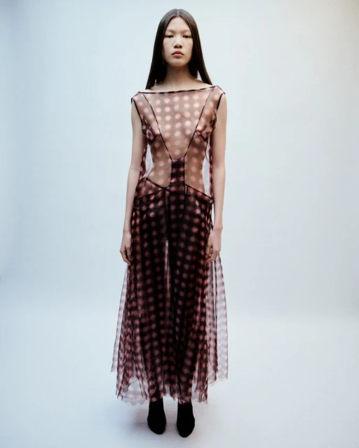 A slender Asian woman in a sheer, dotted dress stands against a white background in a fashion studio. Full-length portrait with soft lighting.