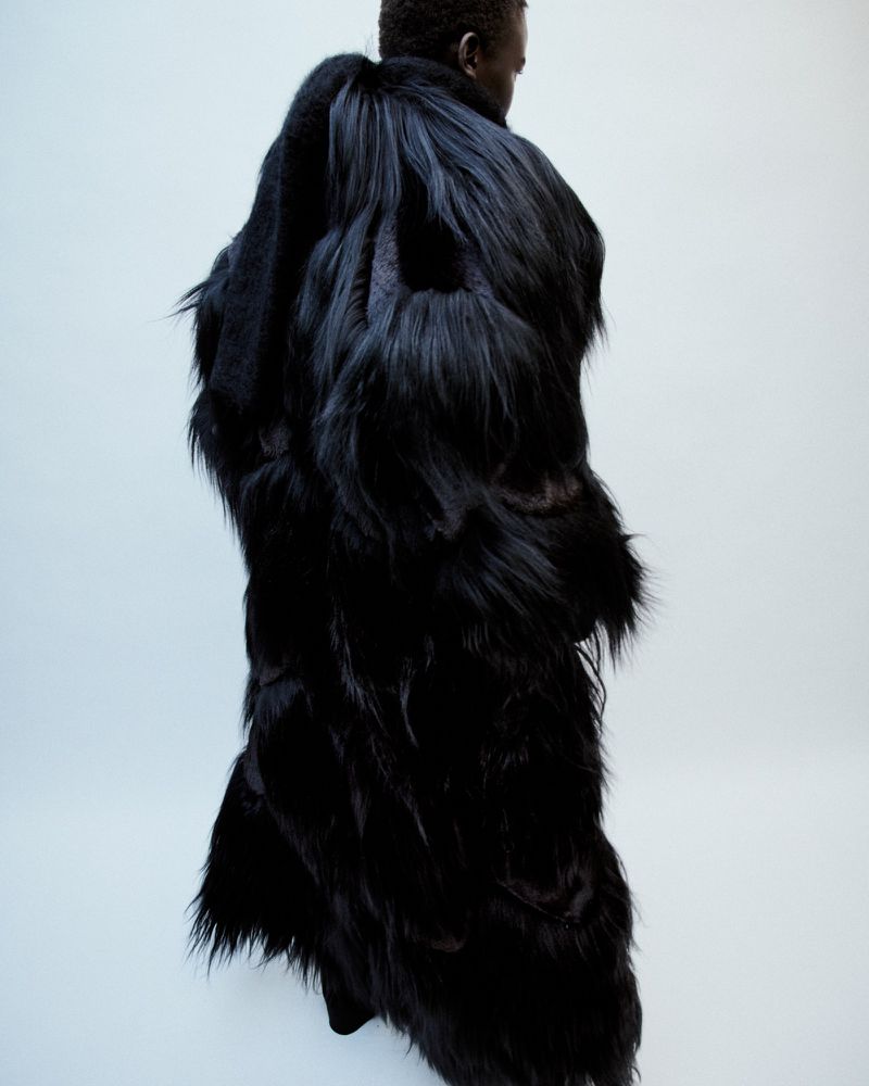Model wearing a voluminous long black textured coat viewed from behind against a plain white background