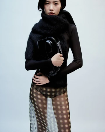 Model in black top and sheer dotted skirt holding a structured handbag against a plain background