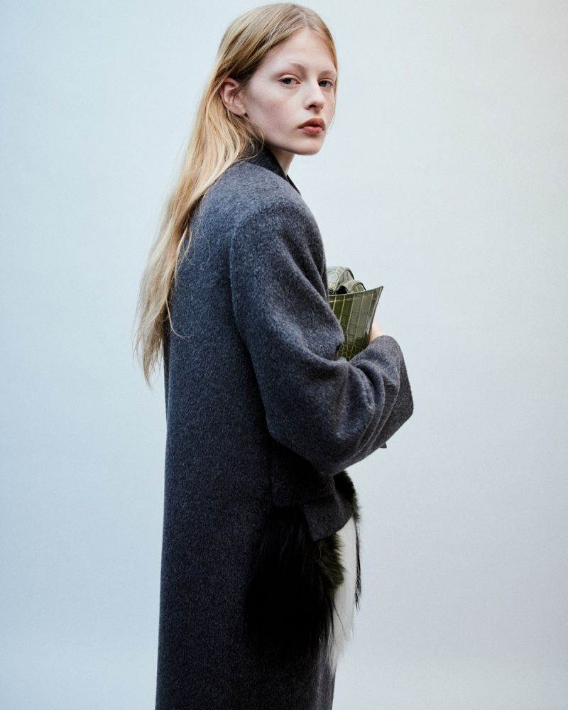 Fashion portrait of a blonde adult woman wearing a long grey coat, and holding an olive green clutch against a plain light background.