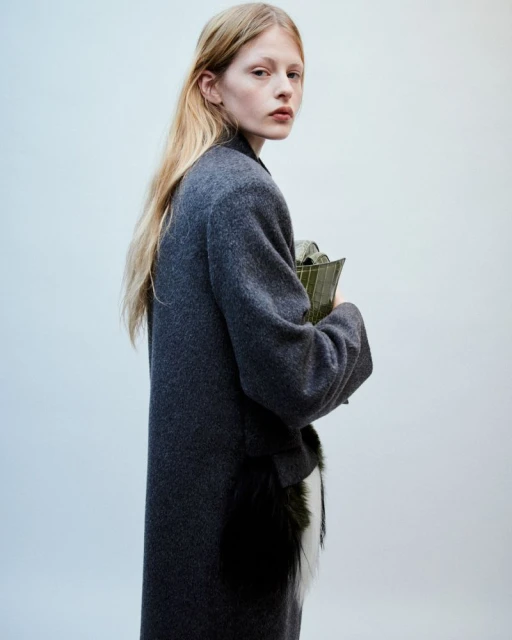 Fashion portrait of a blonde adult woman wearing a long grey coat, and holding an olive green clutch against a plain light background.