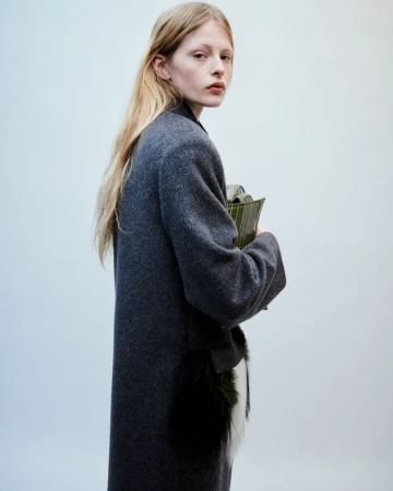 Fashion portrait of a blonde adult woman wearing a long grey coat, and holding an olive green clutch against a plain light background.