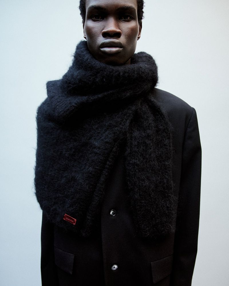 A dark-skinned male fashion model wears a large, fluffy black scarf and black coat against a light neutral background in a studio setting.