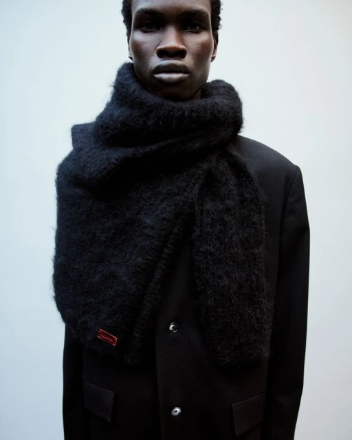 A dark-skinned male fashion model wears a large, fluffy black scarf and black coat against a light neutral background in a studio setting.