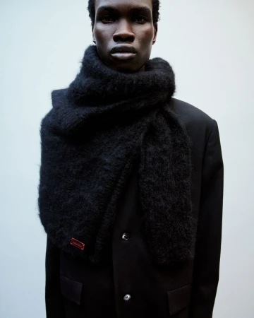 A dark-skinned male fashion model wears a large, fluffy black scarf and black coat against a light neutral background in a studio setting.