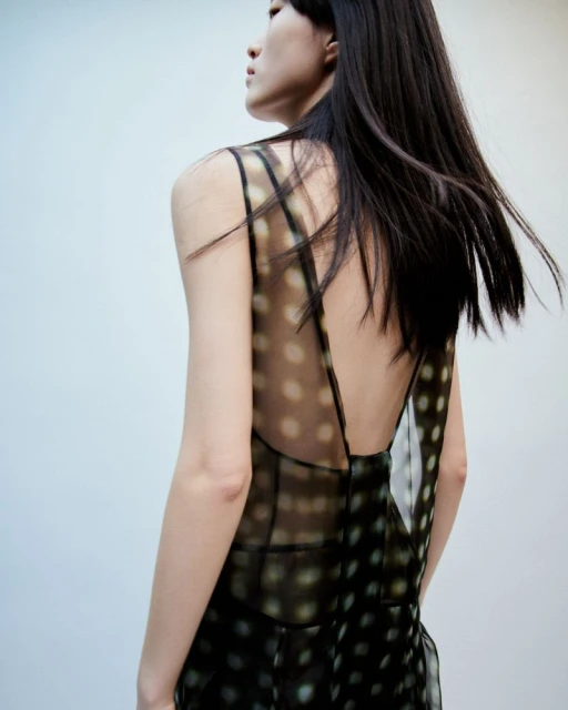 An Asian adult female model showcases a semi-sheer black sleeveless dress with polka dots against a neutral background in a studio shot.