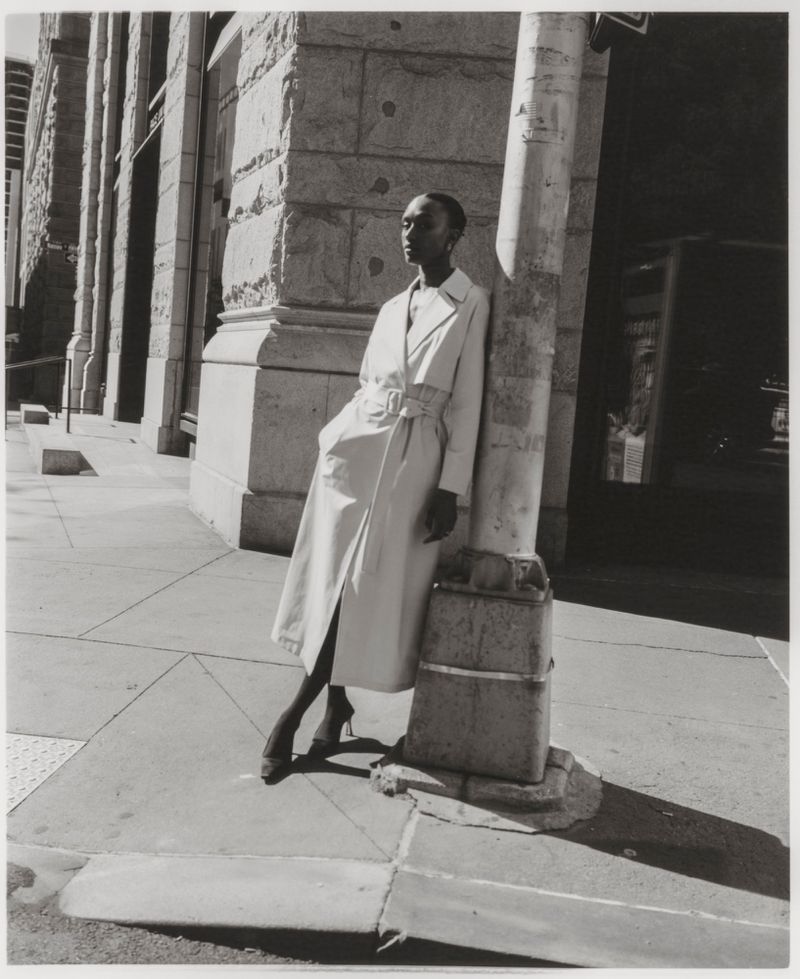 Monochrome outdoor fashion shot of a Black female model in a belted trench coat, leaning against a utility pole on a city street.