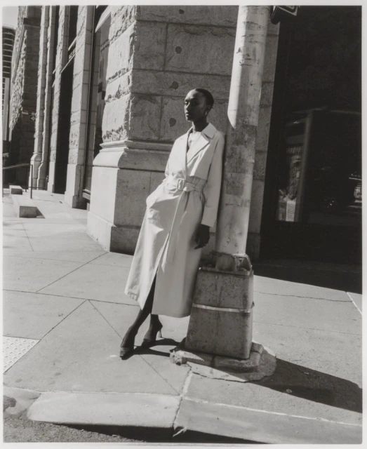 Monochrome outdoor fashion shot of a Black female model in a belted trench coat, leaning against a utility pole on a city street.