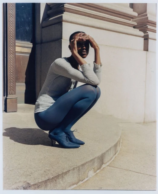 Woman in textured top and blue tights poses on steps of building, shielding her eyes from sun with her hands in urban setting.