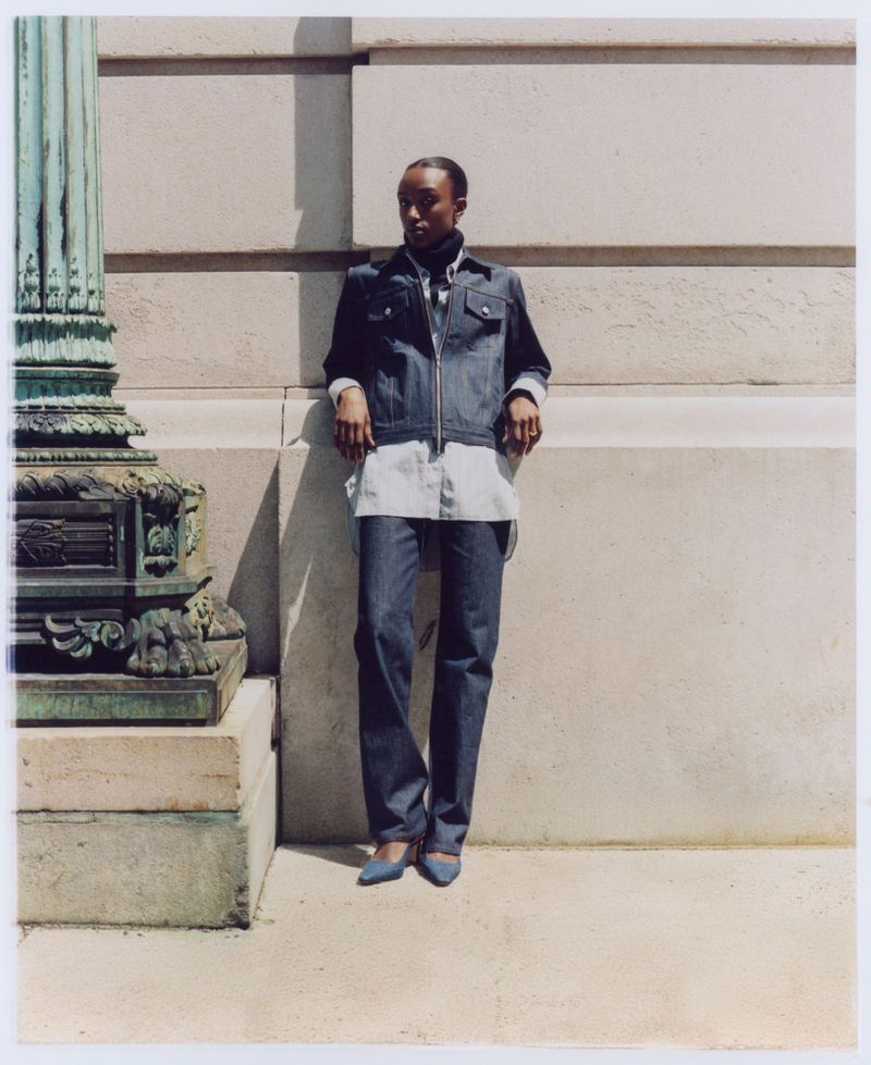 A slender Black woman in a denim jacket, white shirt, and jeans poses confidently against a textured wall near a fluted column.