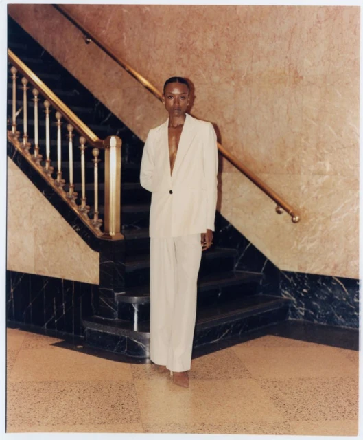 Fashion portrait of a Black female model in a cream-colored suit standing near a staircase with marble walls and black and gold railings.