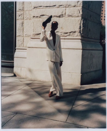 Model Amelie Nsengiyumva wearing an off-white tailored suit and holding a black clutch in sunlight outside a stone building.