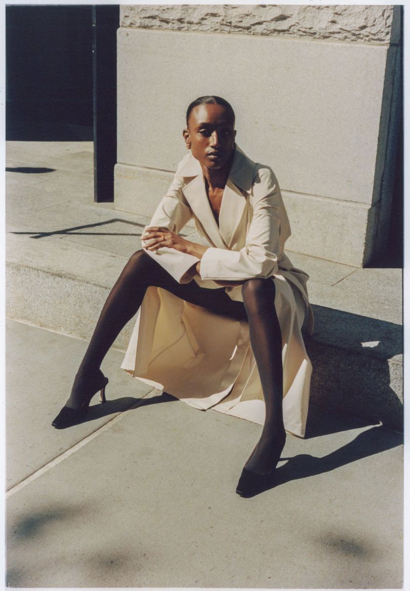 A slender adult wearing a trench coat and heels poses while sitting on concrete steps outside in the sunshine. Style and fashion.