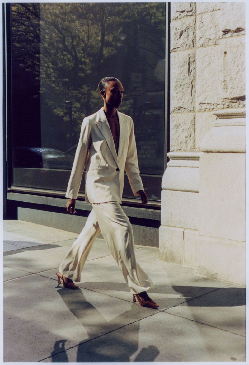 Woman in cream suit walking along city sidewalk near building with window reflection, wearing heels and confident expression.