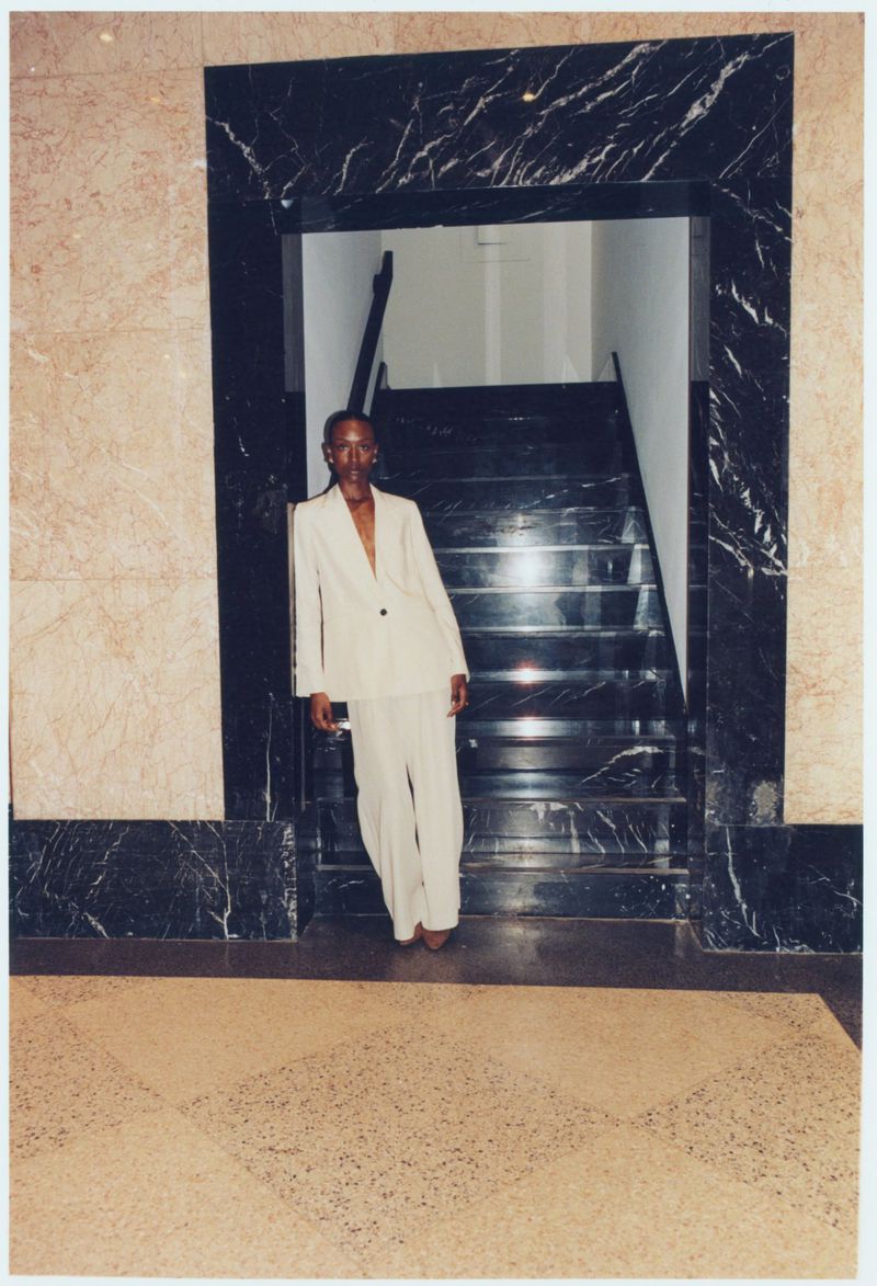 Person in a cream suit stands near a marble staircase and doorway. The scene has natural light, and the neutral color scheme is visible.