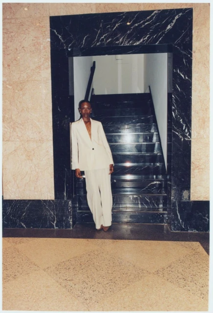 Person in a cream suit stands near a marble staircase and doorway. The scene has natural light, and the neutral color scheme is visible.