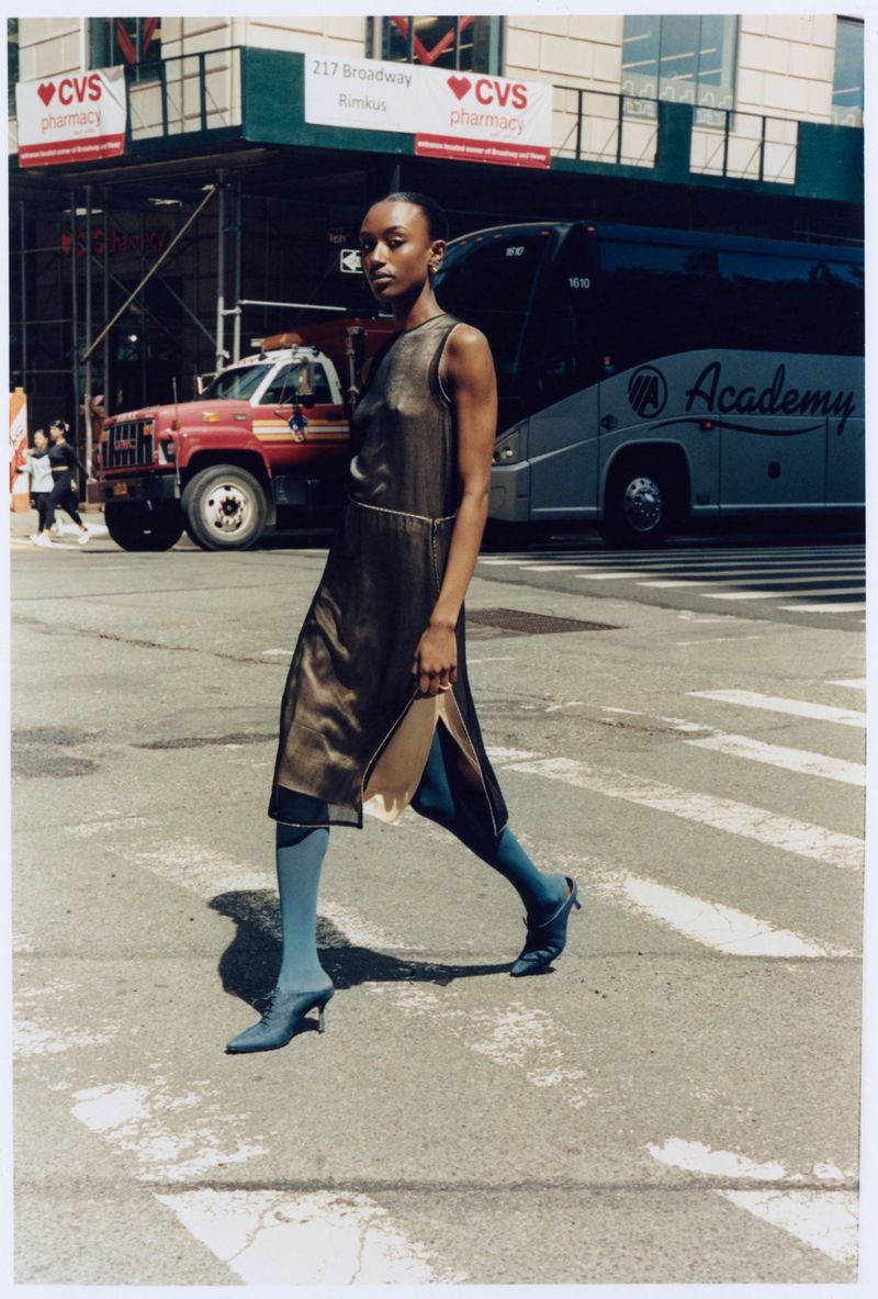 Woman in a sleeveless dress walks across a crosswalk in the city with a fire truck and coach bus in the background on a sunny day.