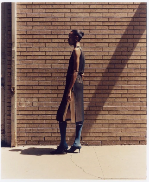A slender Black woman poses in profile against a brick wall wearing a sleeveless dress, blue stockings, and heeled shoes on a sunny day.