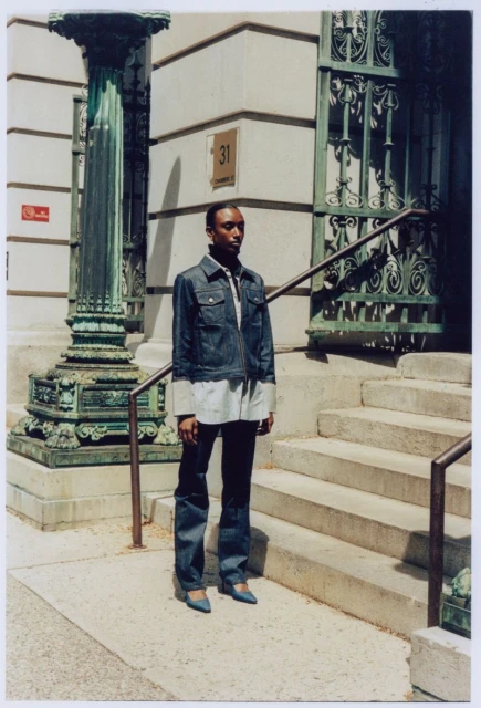 A person stands on concrete steps of a building near an ornate column, wearing a denim jacket, light shirt, dark pants, and blue shoes.