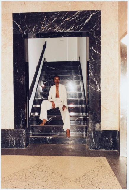 An adult in a light-colored suit sits on marble stairs beneath a dark marble archway, illuminated by natural light, creating an editorial fashion shot.