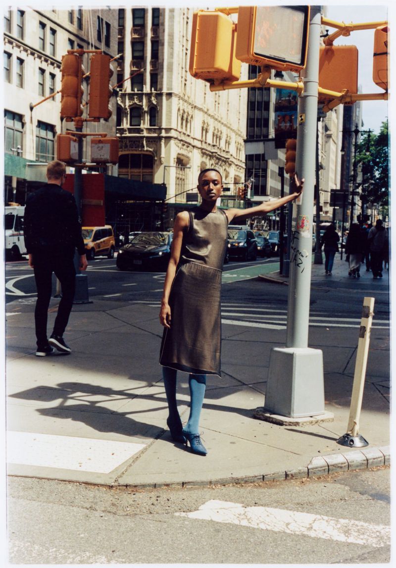A woman in a dress stands on a city street corner next to a traffic light with city buildings in the background on a sunny day.