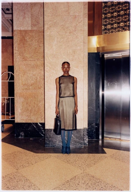 An adult woman poses formally in a lobby wearing a brown dress, blue hosiery and shoes, and carrying a handbag, standing near an elevator.
